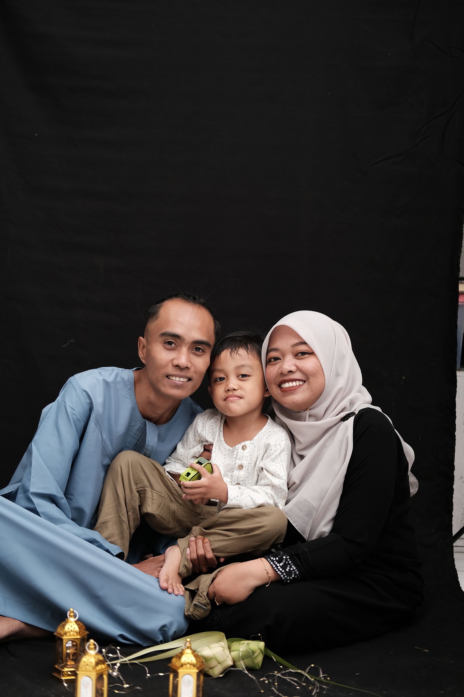 Keluarga Muslim Melayu — bapa berbaju melayu biru, ibu bertudung putih dan anak lelaki kecil — tersenyum bersama tanglung emas dan ketupat, meraikan majlis aqiqah