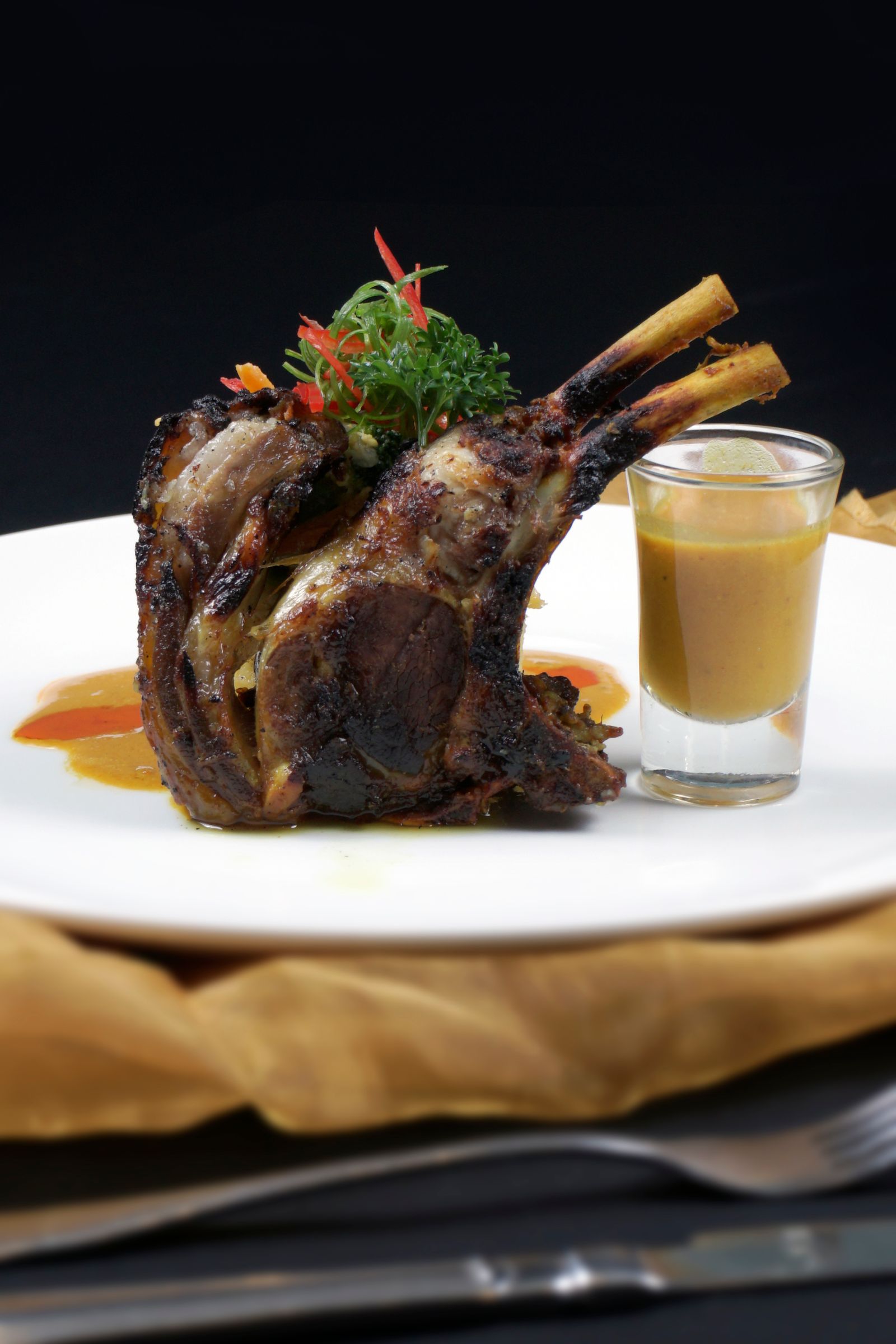 Rak kambing golek dihidang menegak di atas pinggan putih bersama gelas kuah rempah dan hiasan cili