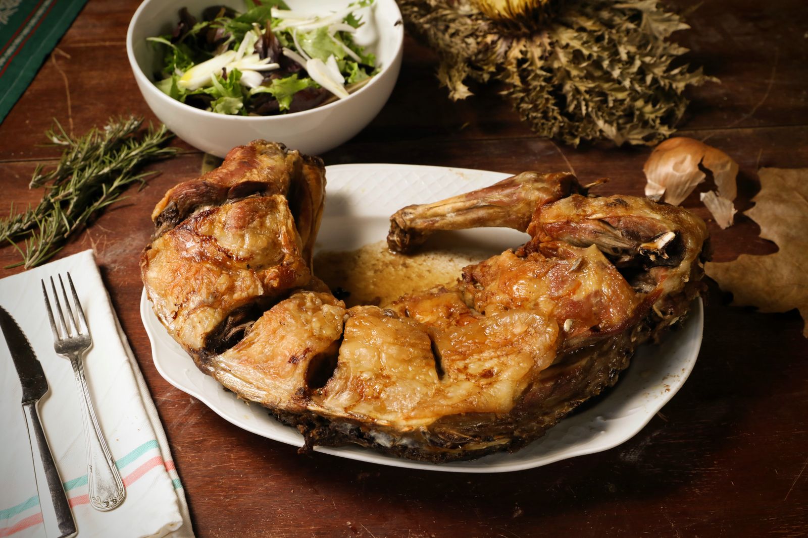Kambing panggang utuh di atas pinggan putih bersama mangkuk salad hijau dan daun rosemary di meja kayu
