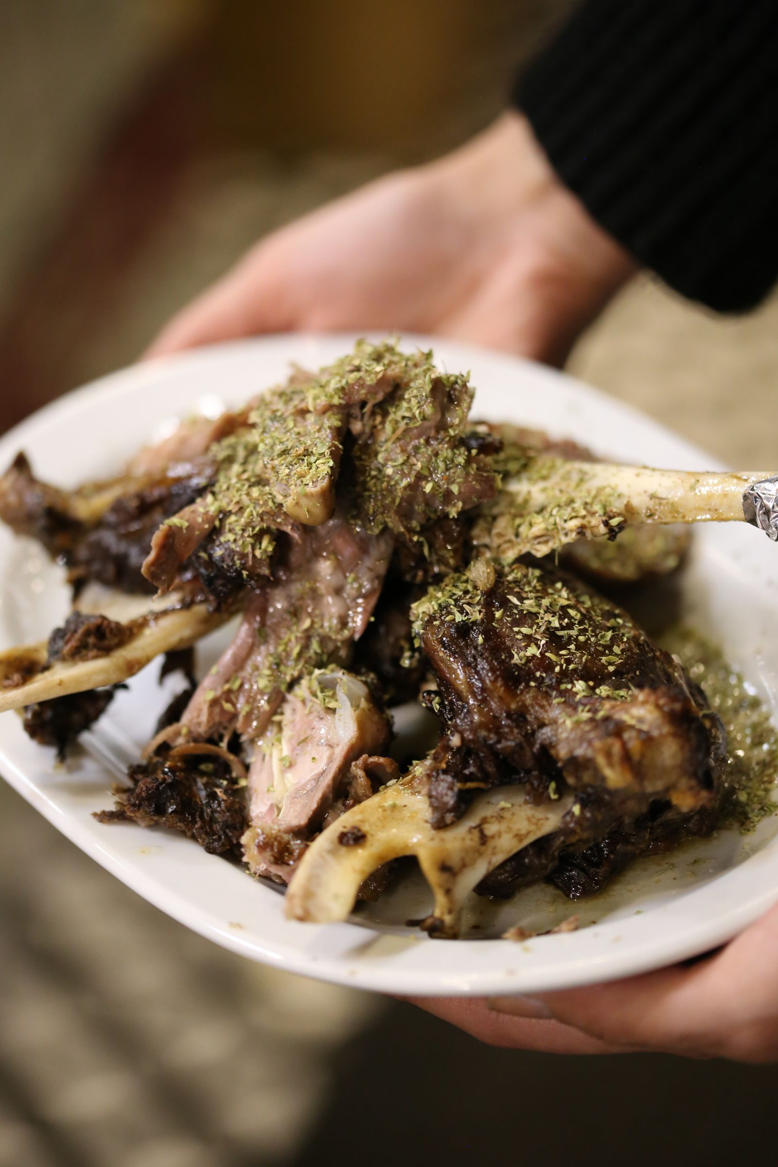 Sepasang tangan menghidang pinggan rusuk kambing panggang bertabur herba kepada tetamu majlis